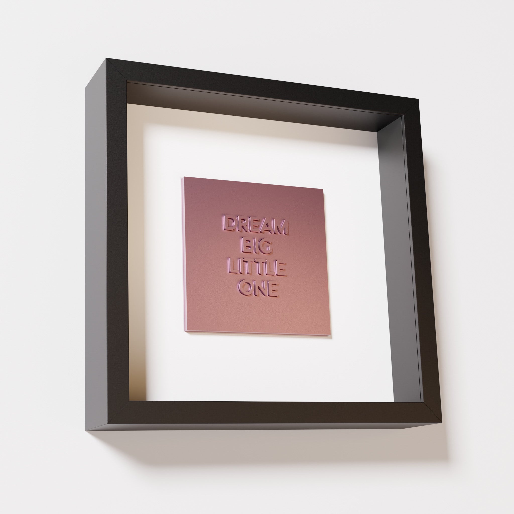 A close-up of a black shadowbox frame containing a metallic rose artwork with the phrase 'Dream big little one,' showcasing bold, raised lettering on a reflective pink surface.