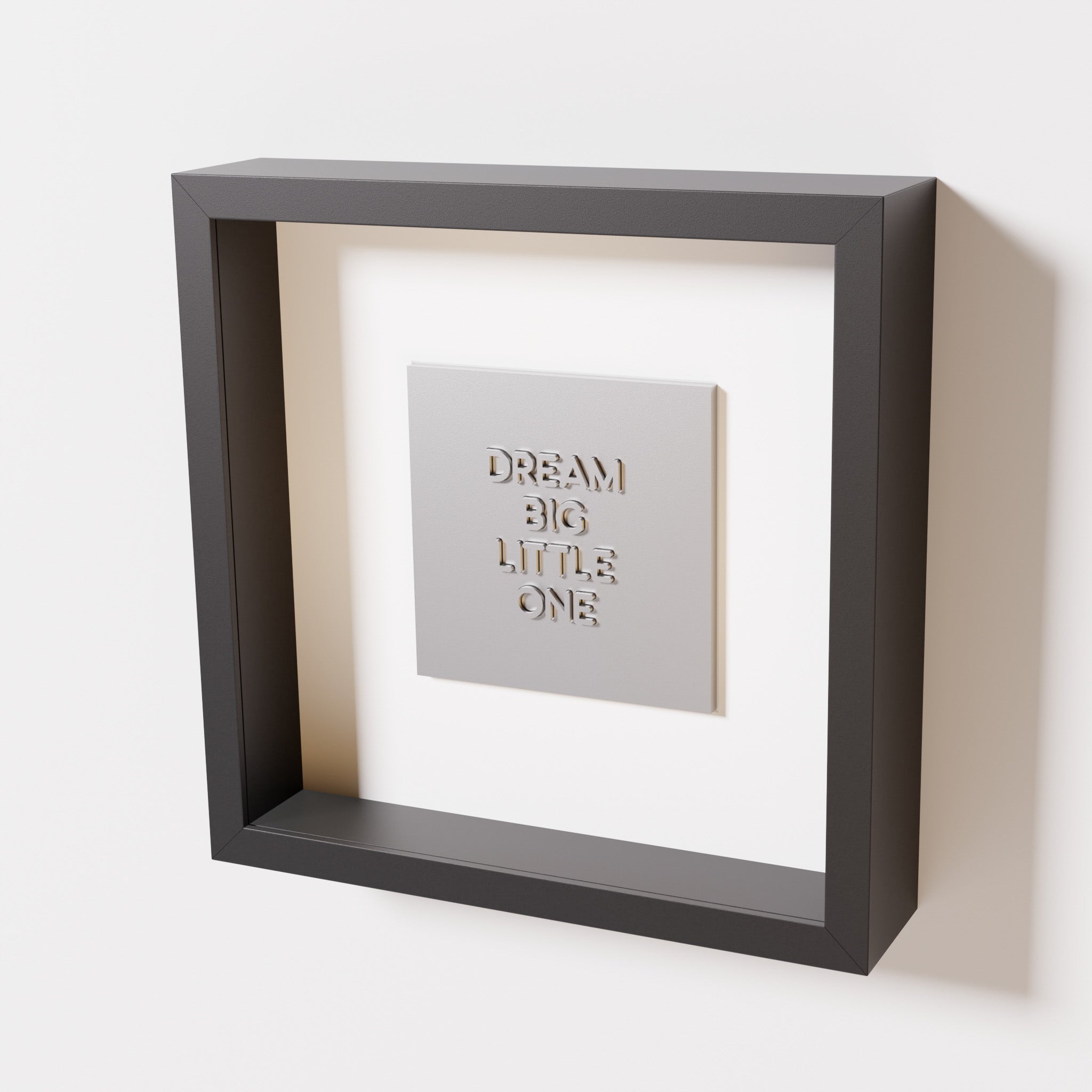 A close-up of a black shadowbox frame containing a chrome silver artwork with the phrase 'Dream big little one,' showcasing bold, raised lettering on a reflective silver surface.