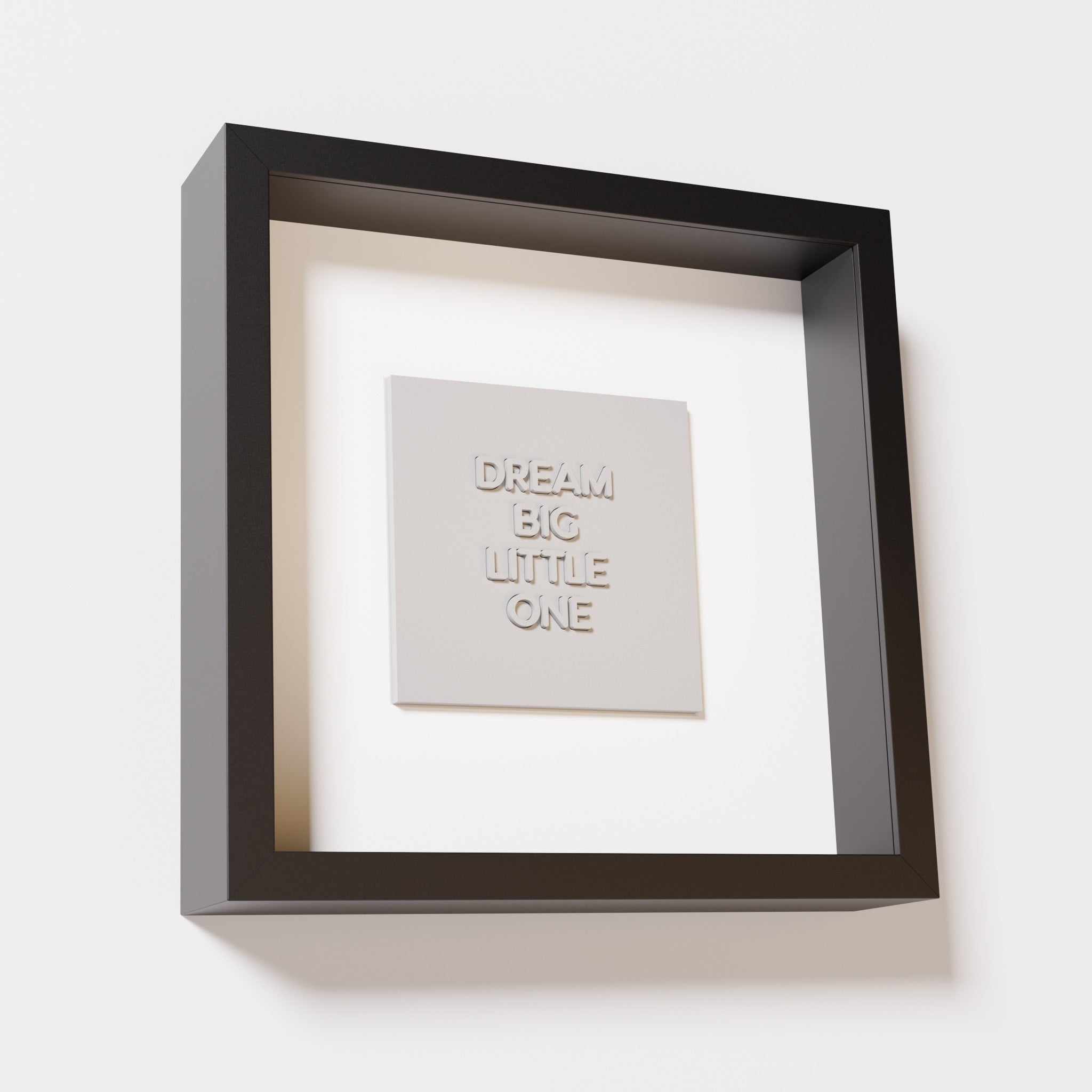 A close-up of a black shadowbox frame containing a marble white artwork with the phrase 'Dream big little one,' showcasing bold, raised lettering on a smooth background.