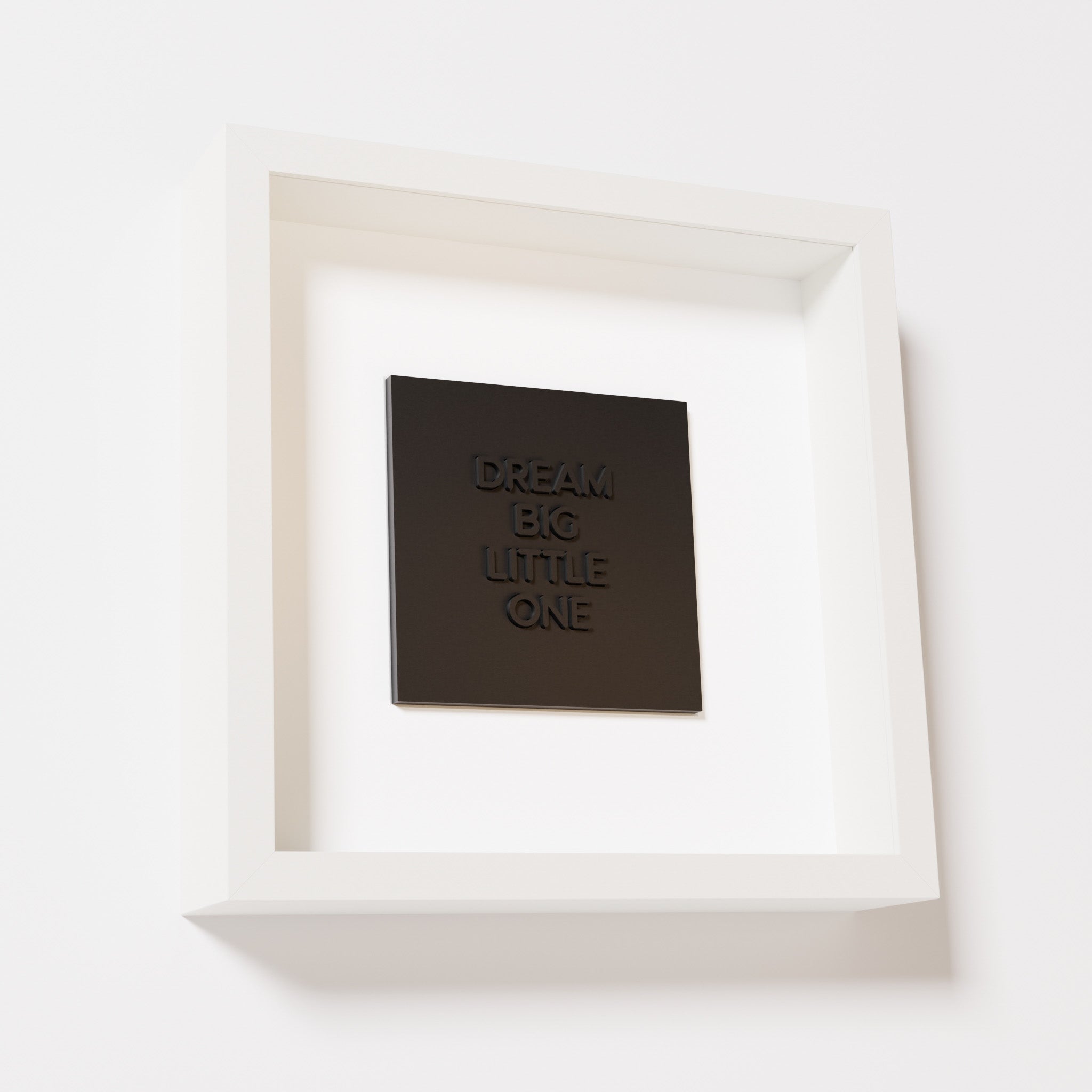 A close-up of a white shadowbox frame containing a matte black artwork with the phrase 'Dream big little one,' showcasing bold, raised lettering on a smooth background.