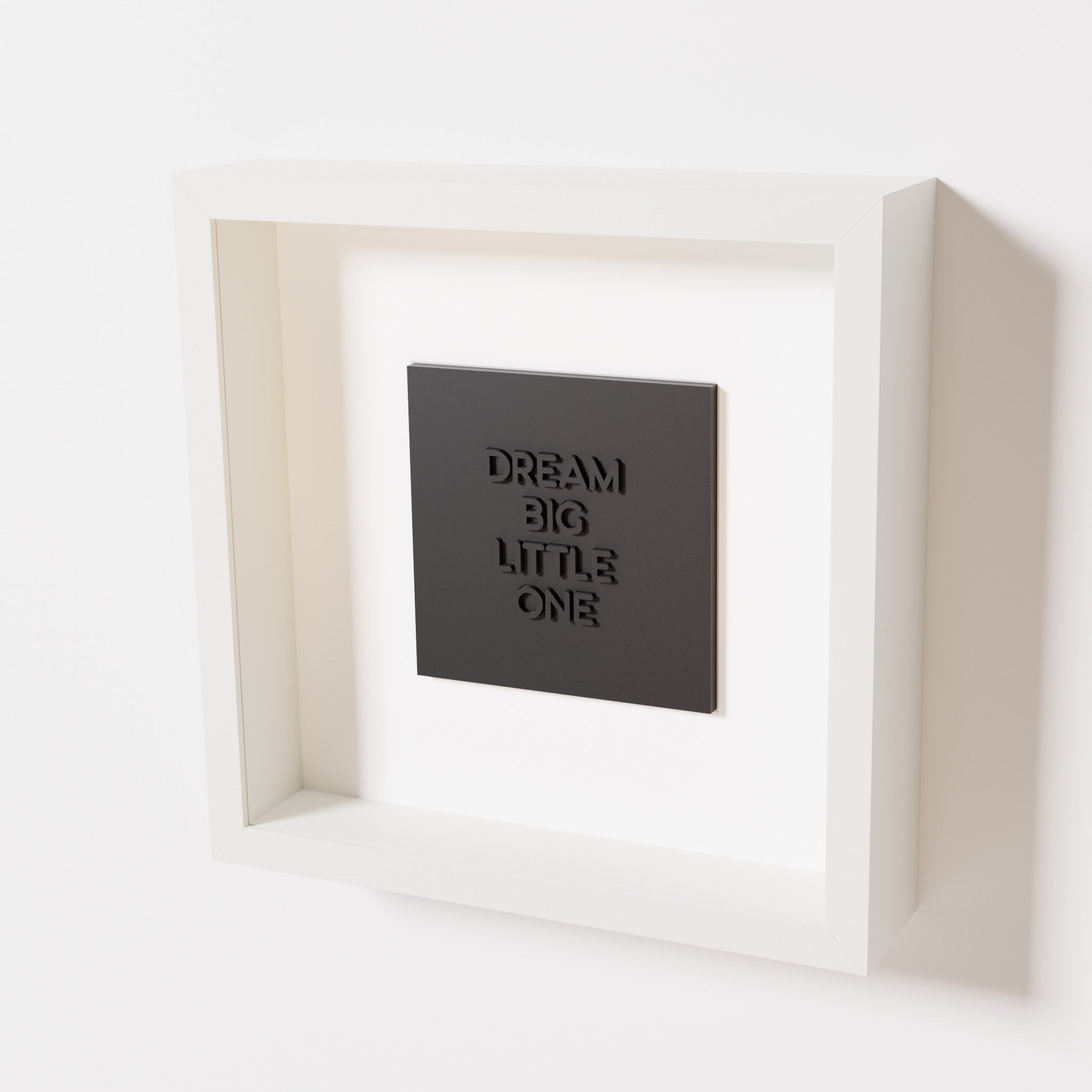 A close-up of a white shadowbox frame containing a matte black artwork with the phrase 'Dream big little one,' showcasing bold, raised lettering on a smooth background.