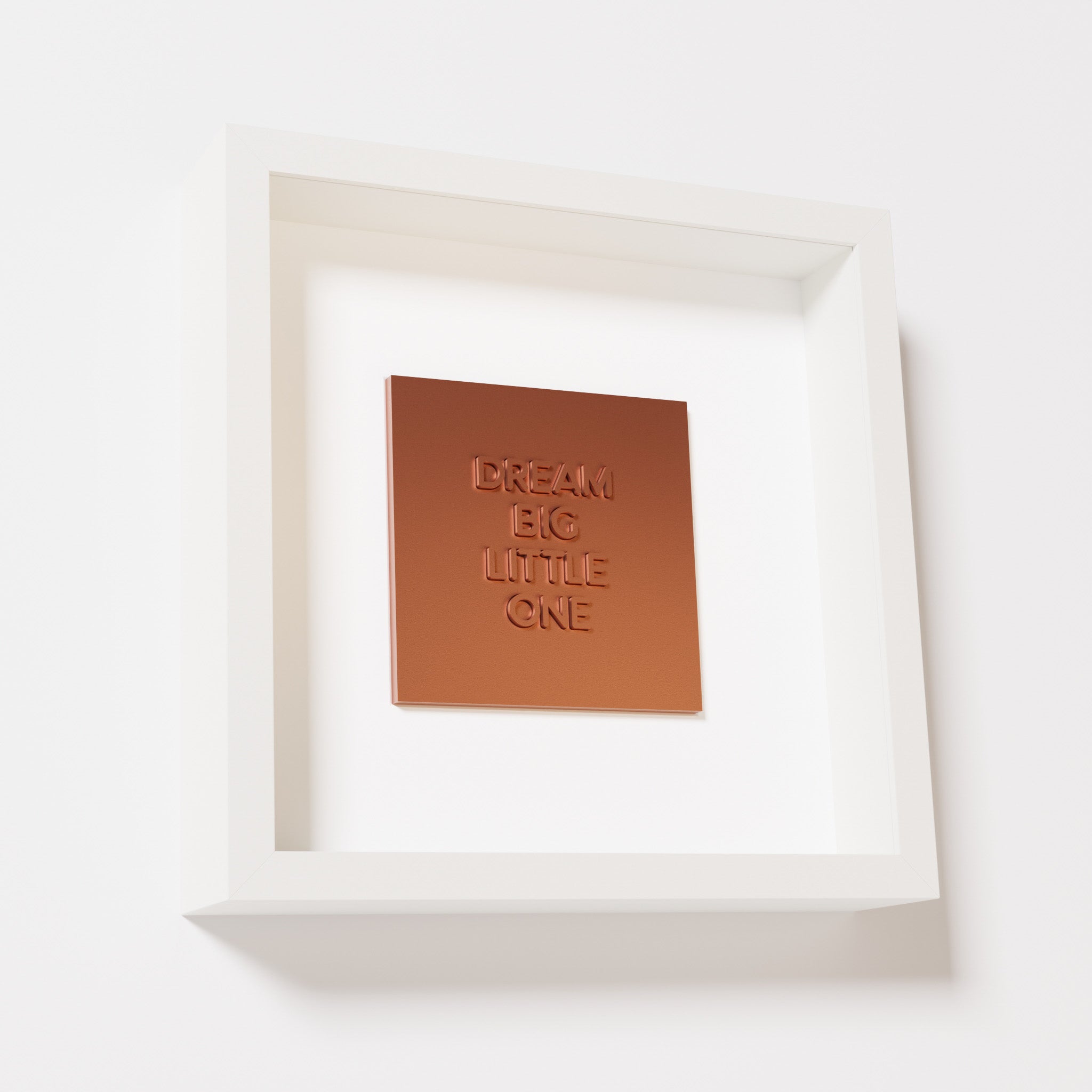 A close-up of a white shadowbox frame containing a chrome copper artwork with the phrase 'Dream big little one,' showcasing bold, raised lettering on a reflective copper surface.