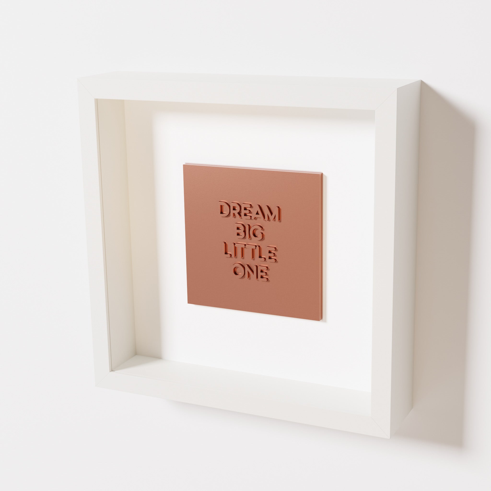 A close-up of a white shadowbox frame containing a chrome copper artwork with the phrase 'Dream big little one,' showcasing bold, raised lettering on a reflective copper surface.