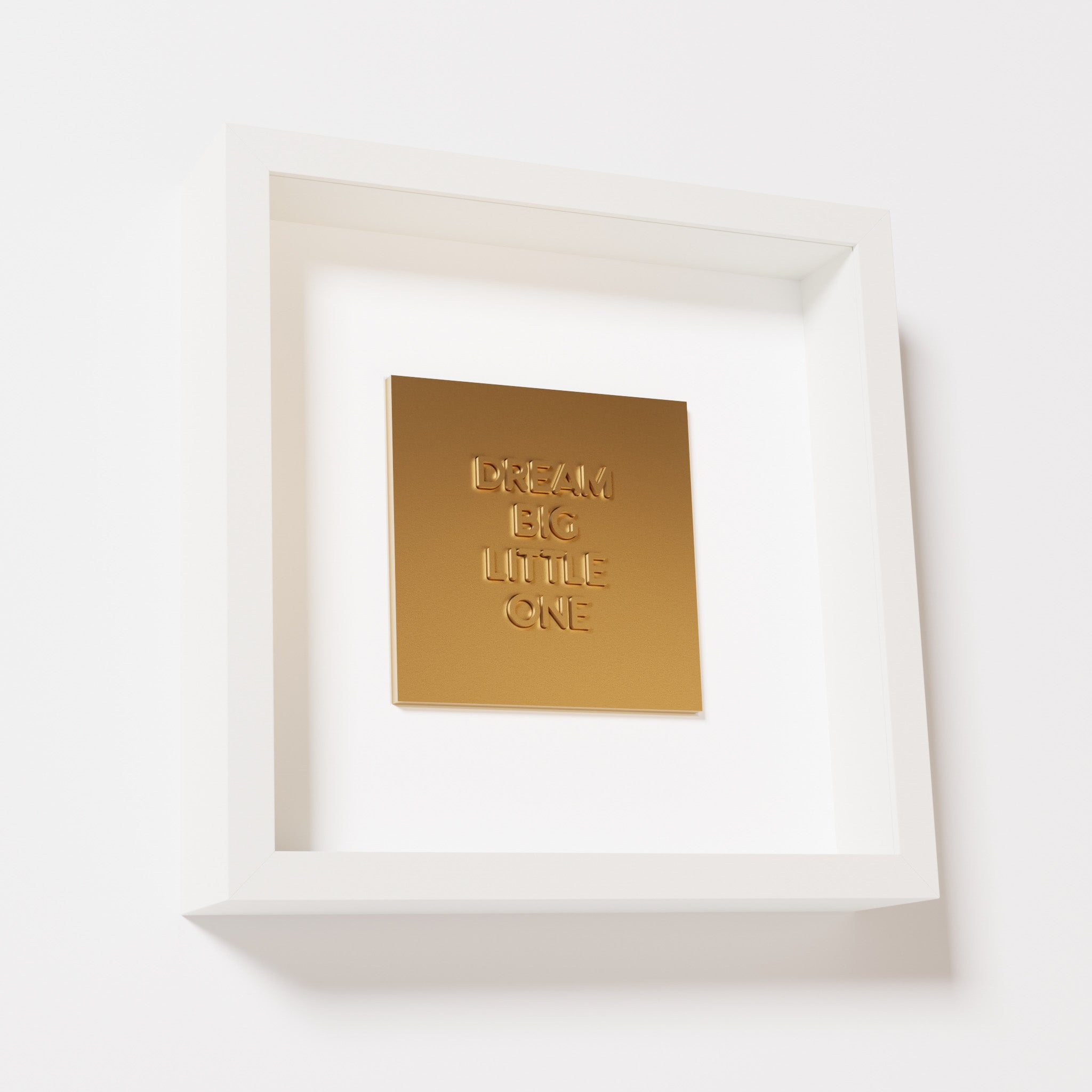 A close-up of a white shadowbox frame containing a chrome gold artwork with the phrase 'Dream big little one,' showcasing bold, raised lettering on a reflective golden surface.