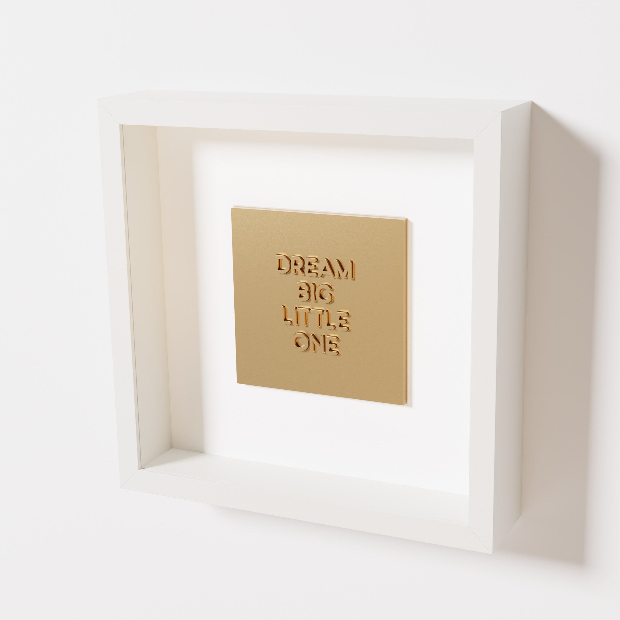 A close-up of a white shadowbox frame containing a chrome gold artwork with the phrase 'Dream big little one,' showcasing bold, raised lettering on a reflective golden surface.