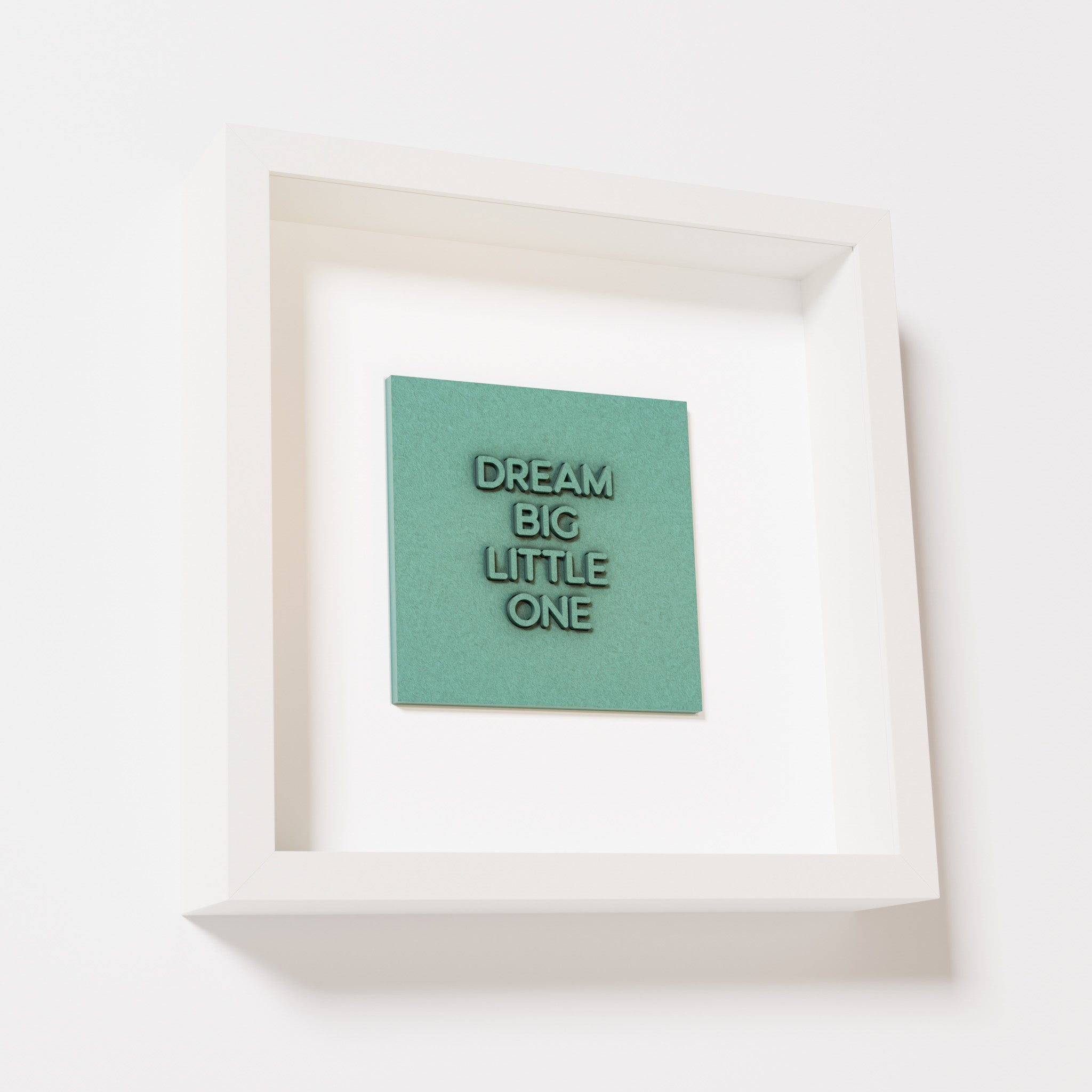 A close-up of a white shadowbox frame containing an old patina artwork with the phrase 'Dream big little one,' showcasing bold, raised lettering on a textured green surface.