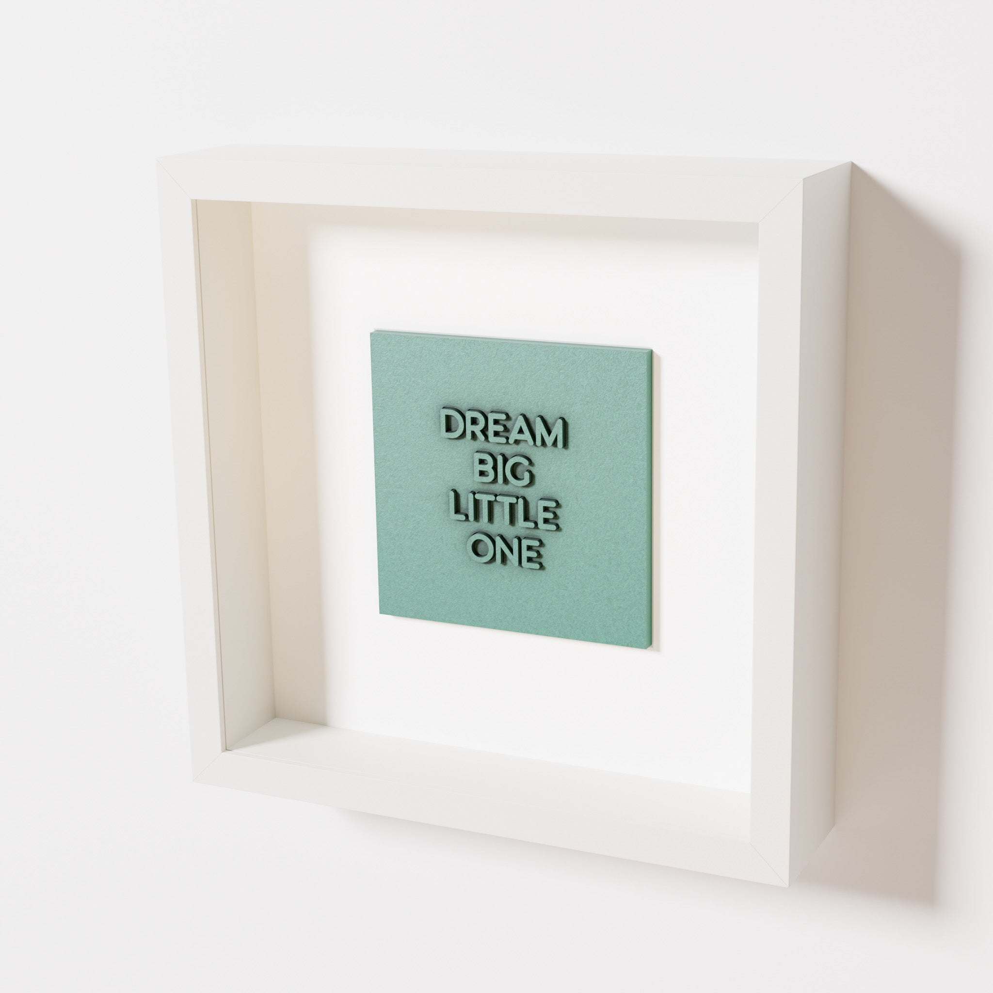 A close-up of a white shadowbox frame containing an old patina artwork with the phrase 'Dream big little one,' showcasing bold, raised lettering on a textured green surface.