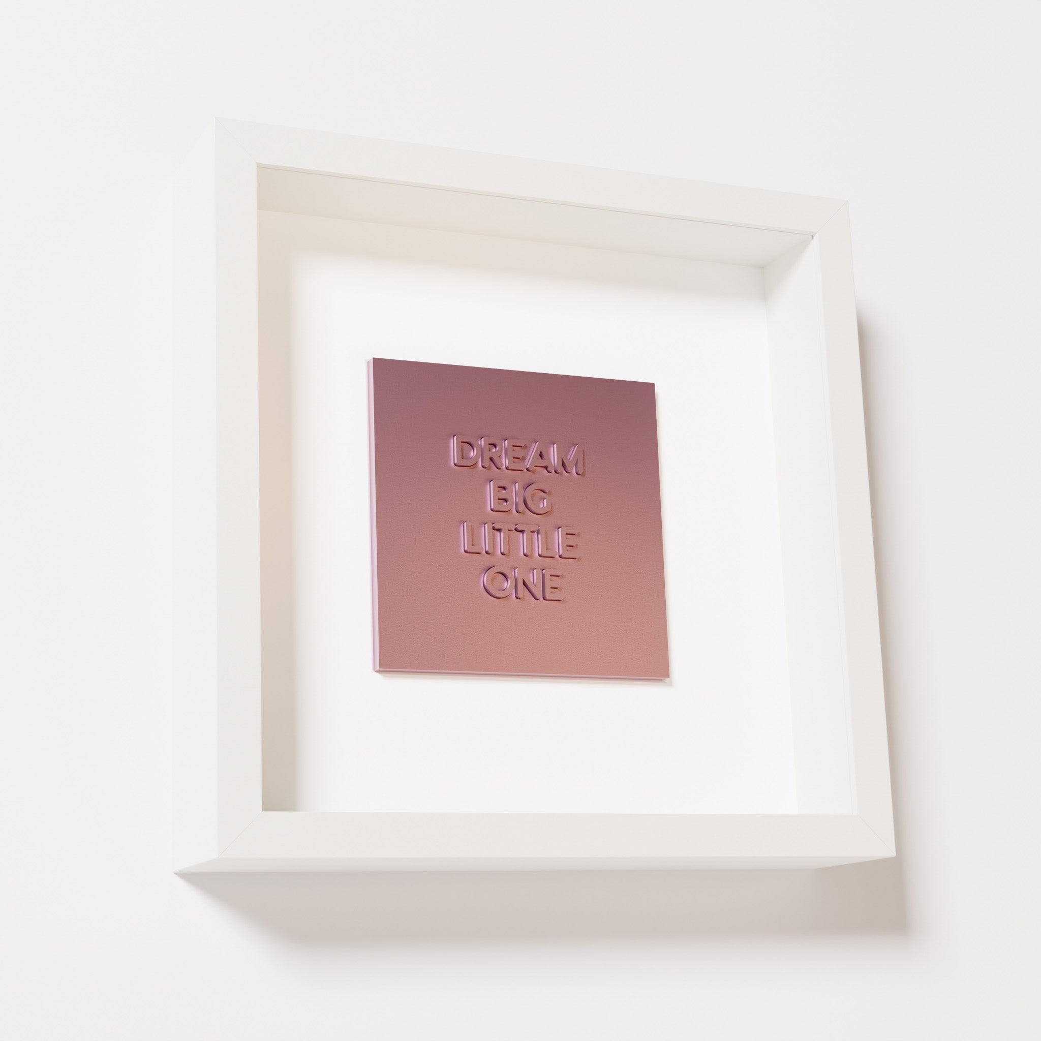 A close-up of a white shadowbox frame containing a metallic rose artwork with the phrase 'Dream big little one,' showcasing bold, raised lettering on a reflective pink surface.