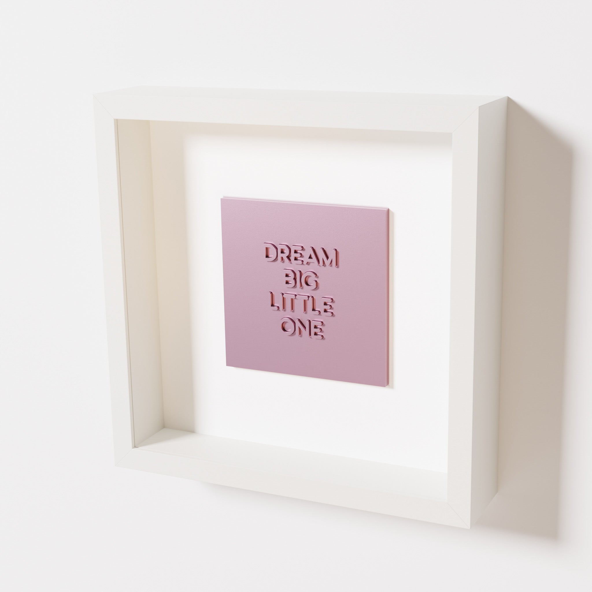 A close-up of a white shadowbox frame containing a metallic rose artwork with the phrase 'Dream big little one,' showcasing bold, raised lettering on a reflective pink surface.