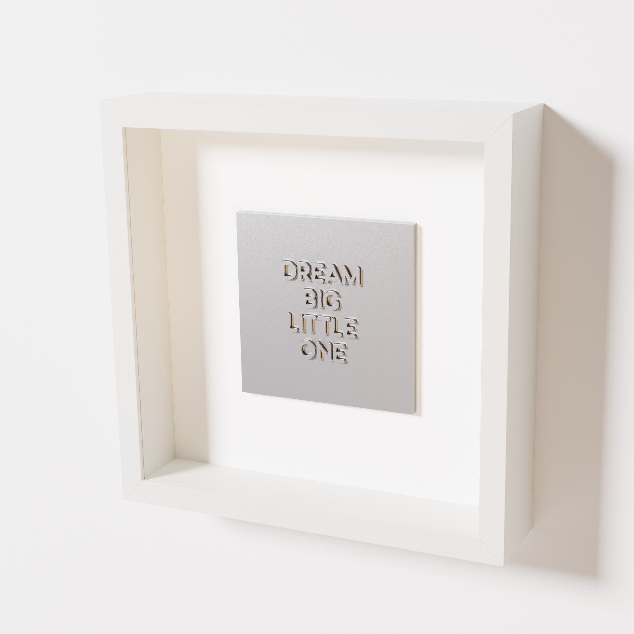 A close-up of a white shadowbox frame containing a chrome silver artwork with the phrase 'Dream big little one,' showcasing bold, raised lettering on a reflective silver surface.