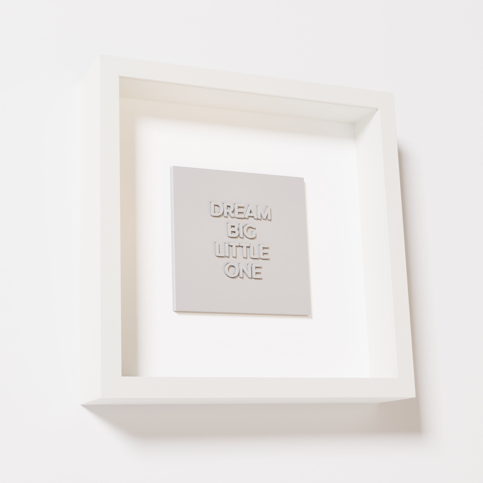 A close-up of a white shadowbox frame containing a marble white artwork with the phrase 'Dream big little one,' showcasing bold, raised lettering on a smooth background.