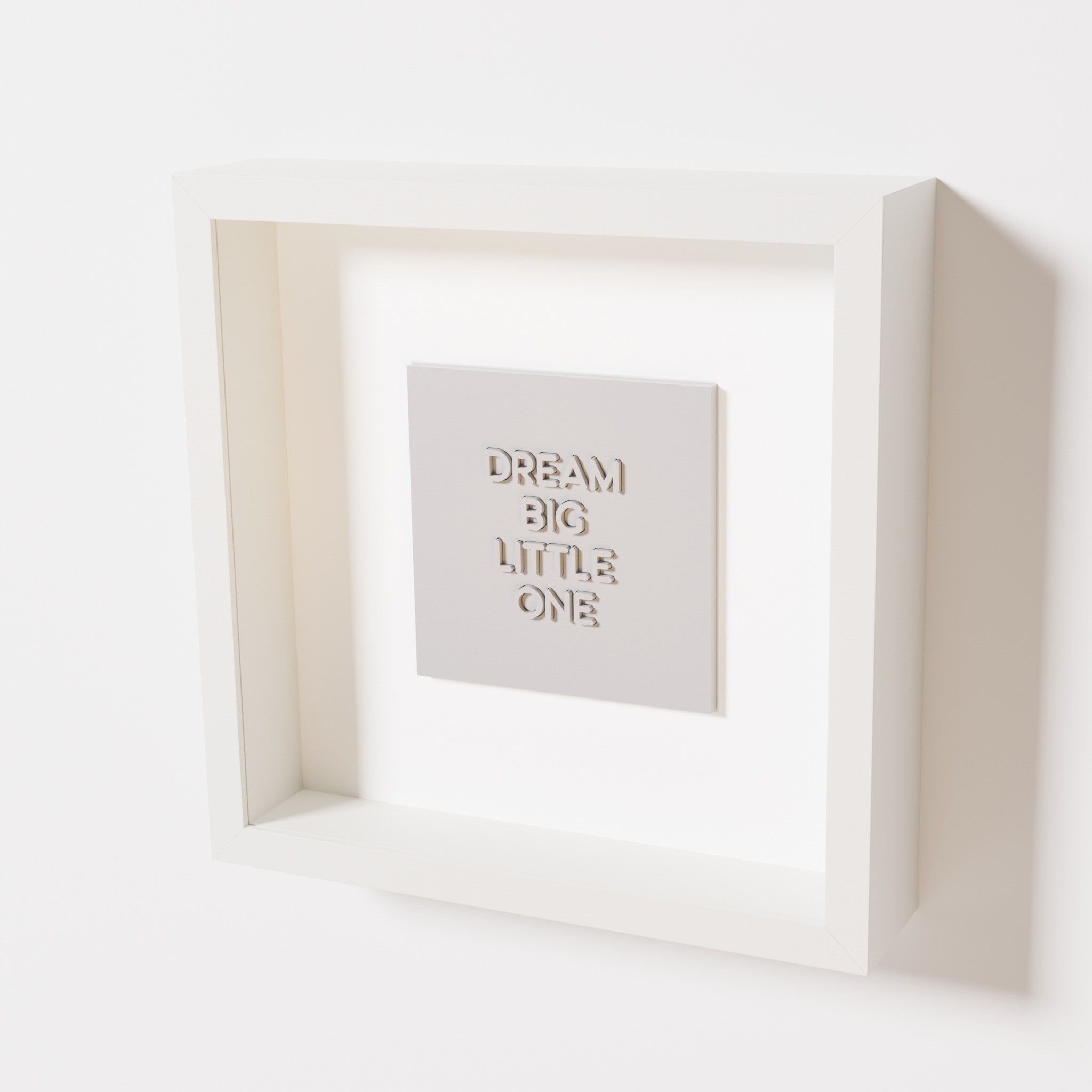 A close-up of a white shadowbox frame containing a marble white artwork with the phrase 'Dream big little one,' showcasing bold, raised lettering on a smooth background.