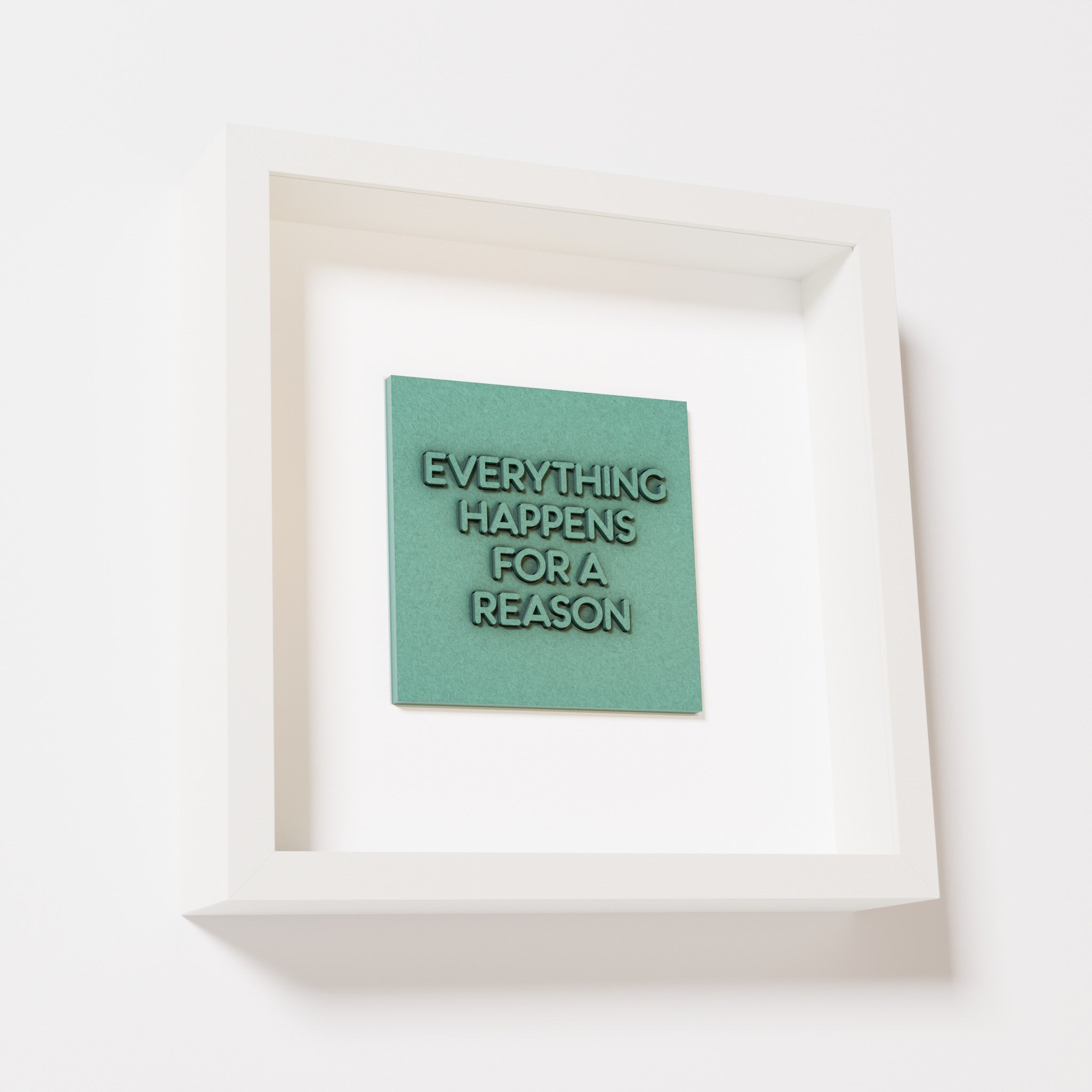 A close-up of a white shadowbox frame containing an old patina artwork featuring the thoughtful phrase 'Everything Happens for a Reason,' emphasizing its textured green finish and modern design.