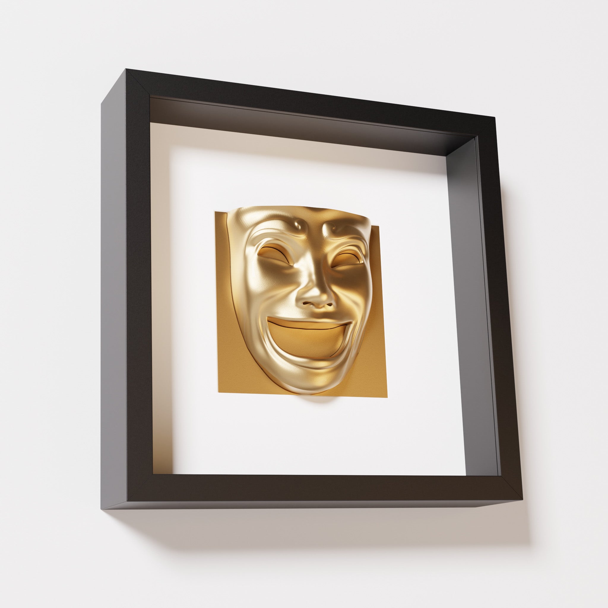 A close-up of a black shadowbox frame containing a chrome gold sculpture of 'Joy,' featuring a theatrical mask with a wide smile and expressive features, evoking happiness and positivity.