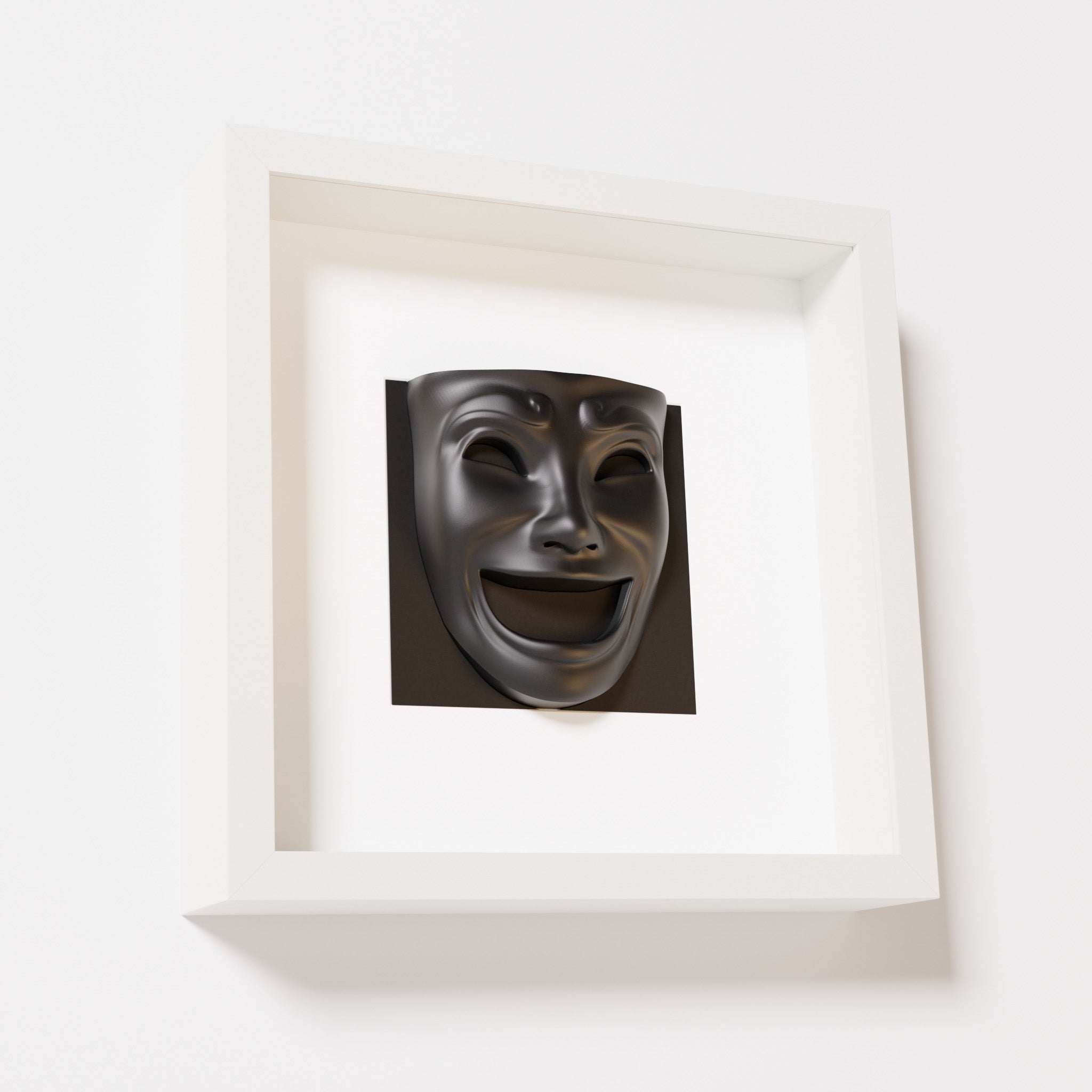 A close-up of a white shadowbox frame containing a matte black sculpture of 'Joy,' featuring a theatrical mask with a wide smile and expressive features, evoking happiness and positivity.