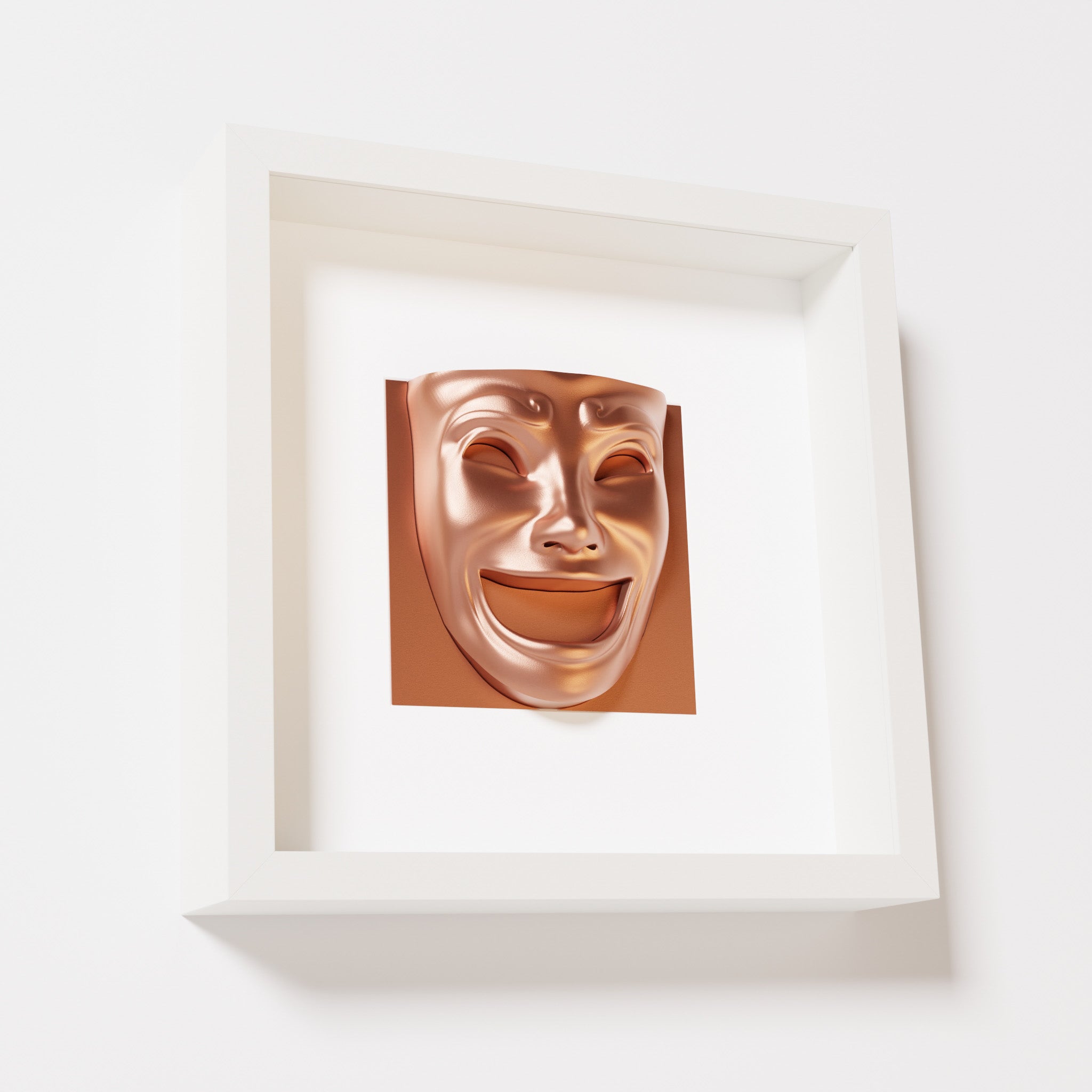 A close-up of a white shadowbox frame containing a chrome copper sculpture of 'Joy,' featuring a theatrical mask with a wide smile and expressive features, evoking happiness and positivity.