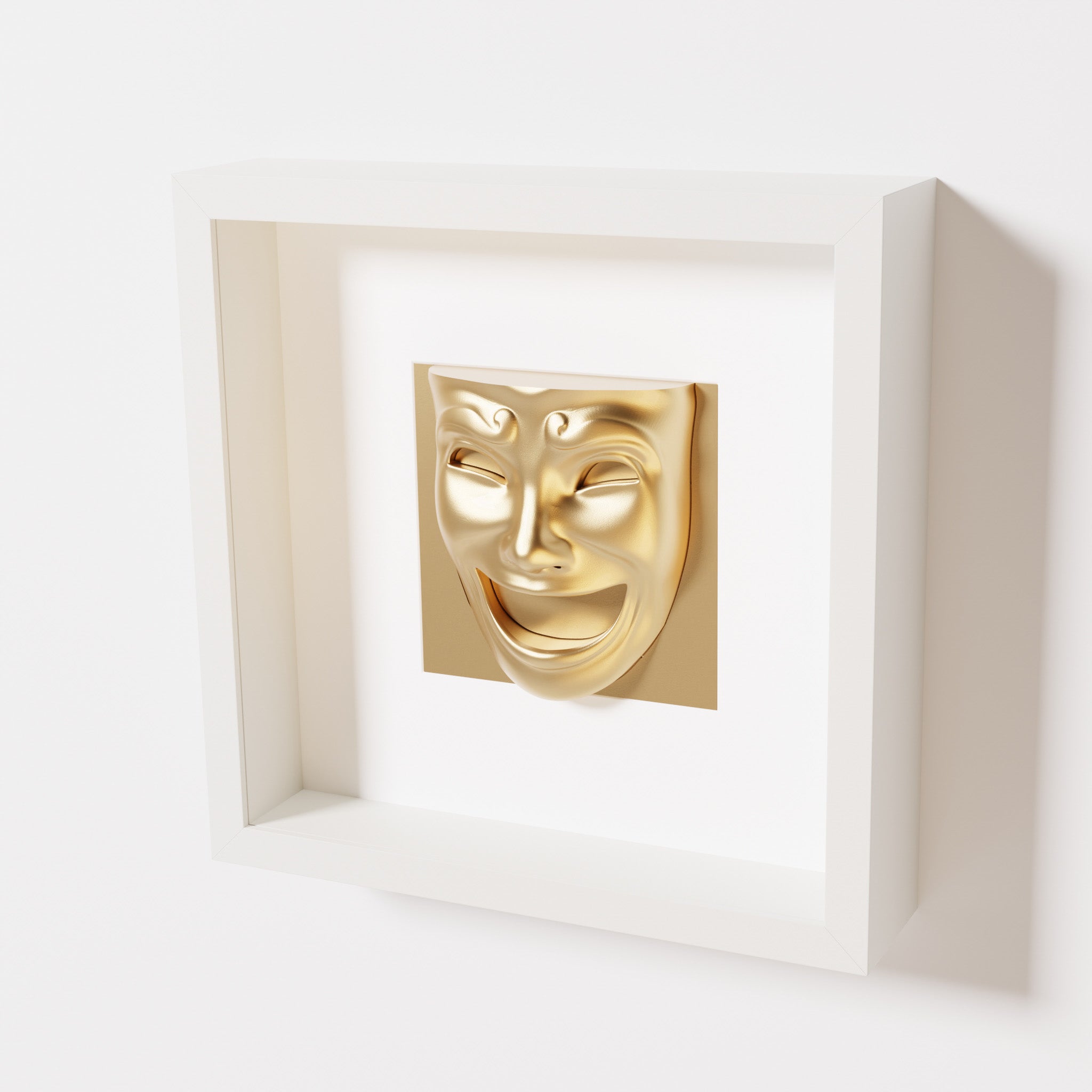 A close-up of a white shadowbox frame containing a chrome gold sculpture of 'Joy,' featuring a theatrical mask with a wide smile and expressive features, evoking happiness and positivity.