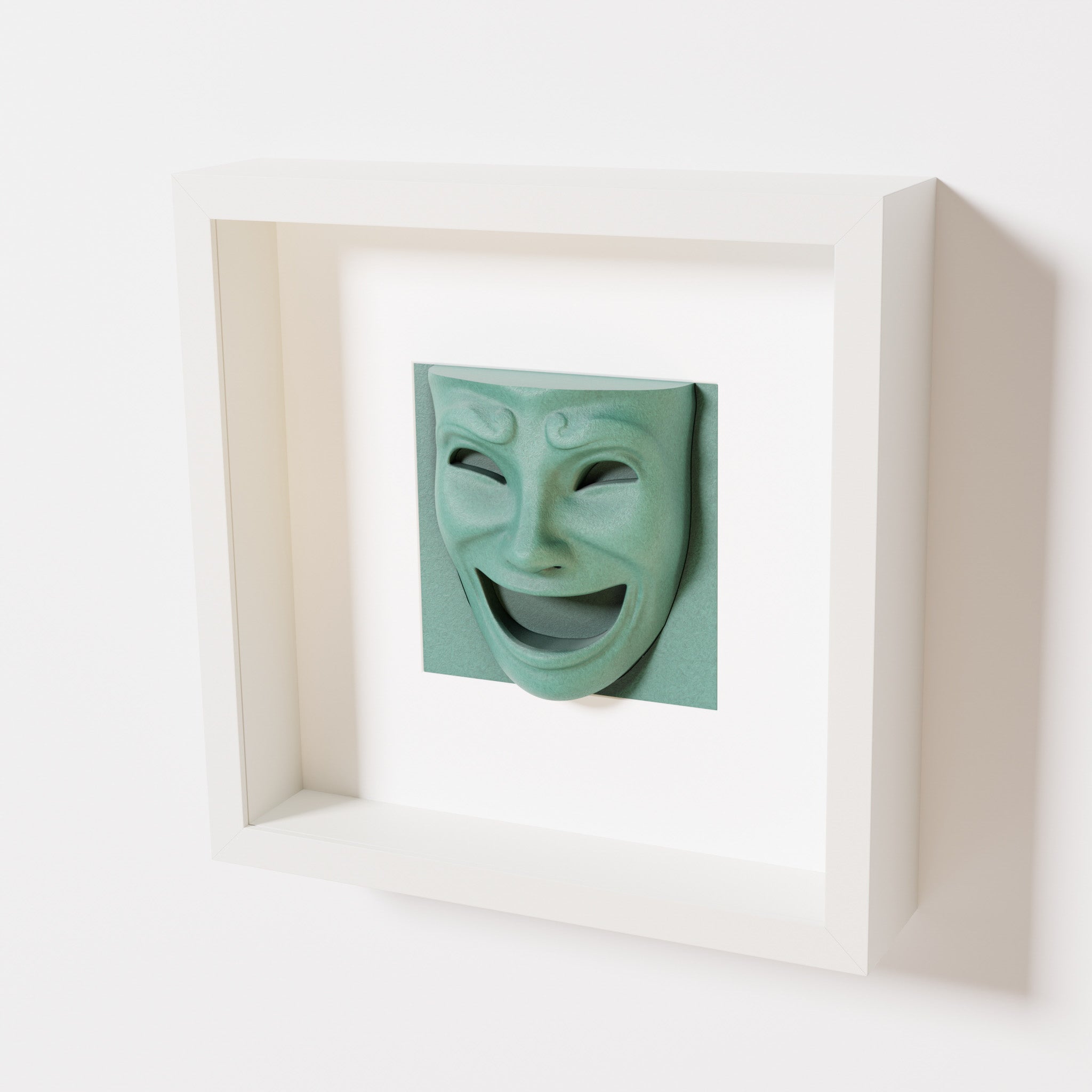 A close-up of a white shadowbox frame containing an old patina sculpture of 'Joy,' featuring a theatrical mask with a wide smile and expressive features, evoking happiness and positivity.