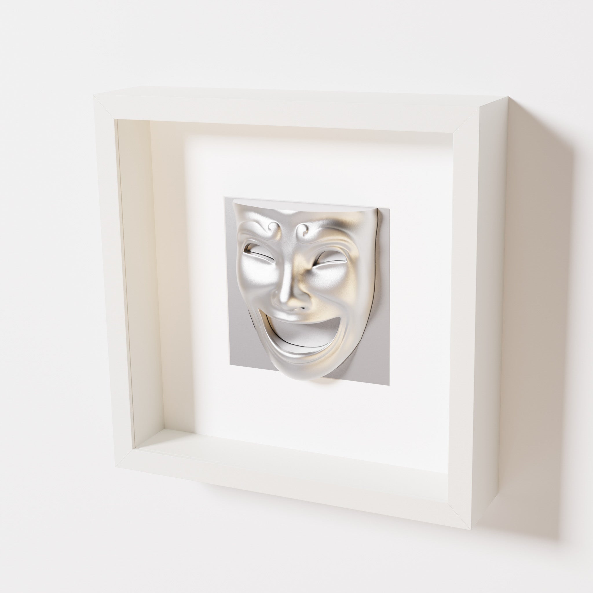 A close-up of a white shadowbox frame containing a chrome silver sculpture of 'Joy,' featuring a theatrical mask with a wide smile and expressive features, evoking happiness and positivity.