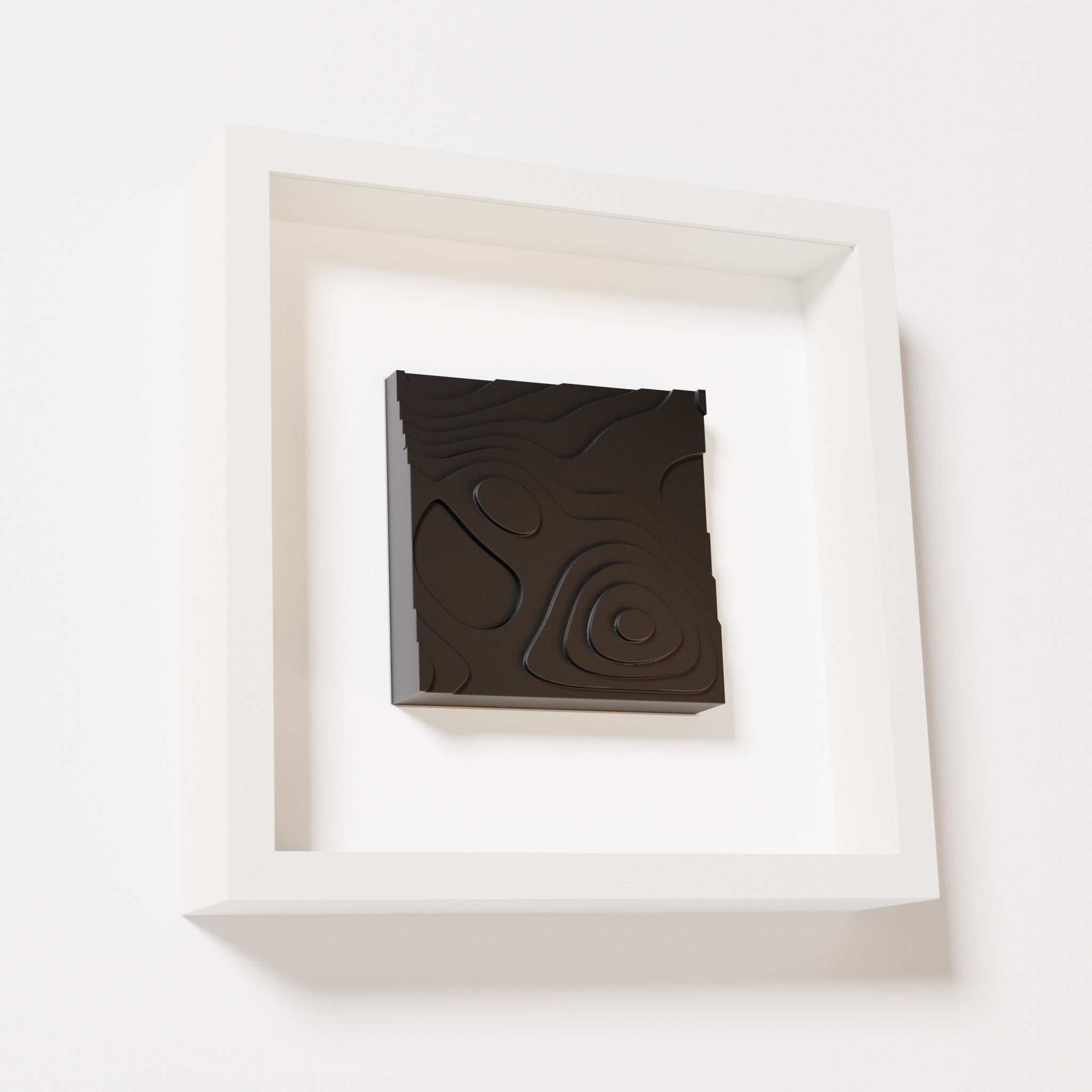 A close-up of a white shadowbox frame containing a matte black artwork depicting layered contour lines inspired by geographic elevation levels, showcasing intricate and geometric details.