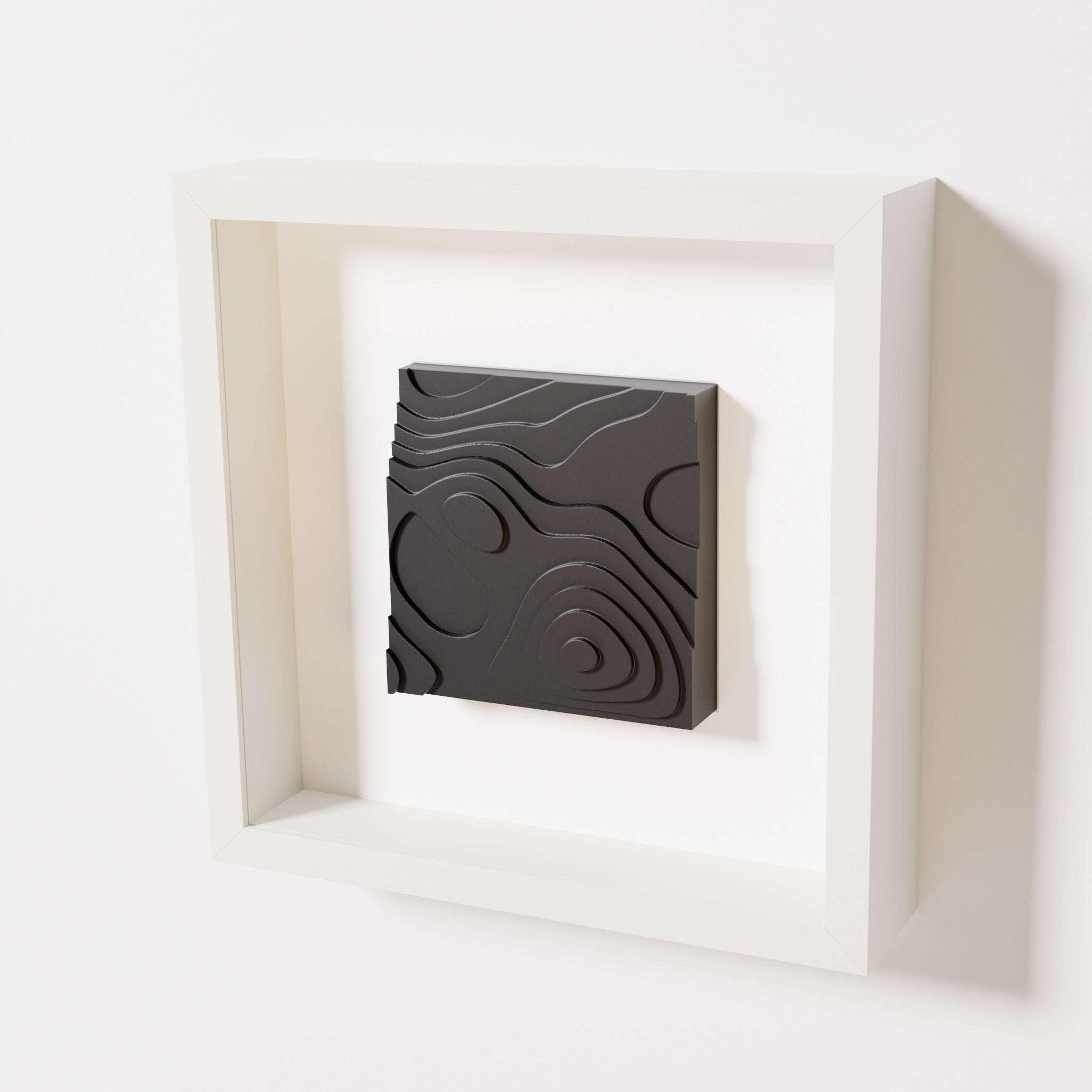 A close-up of a white shadowbox frame containing a matte black artwork depicting layered contour lines inspired by geographic elevation levels, showcasing intricate and geometric details.