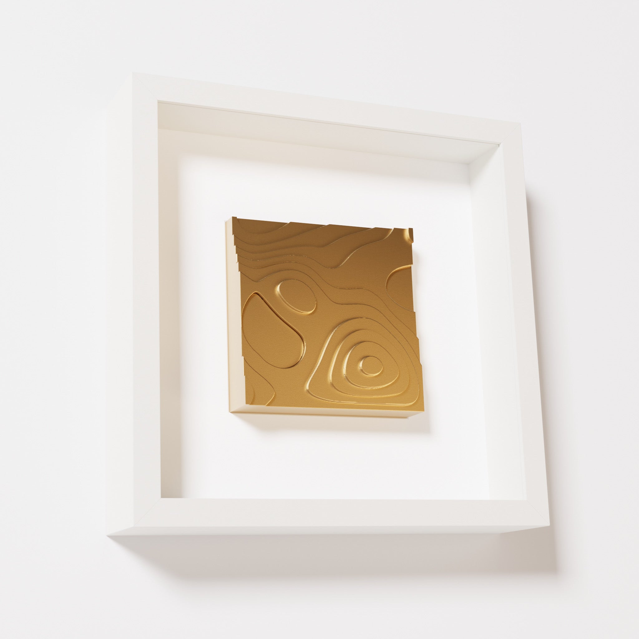 A close-up of a white shadowbox frame containing a chrome gold artwork depicting layered contour lines inspired by geographic elevation levels, showcasing reflective golden details.