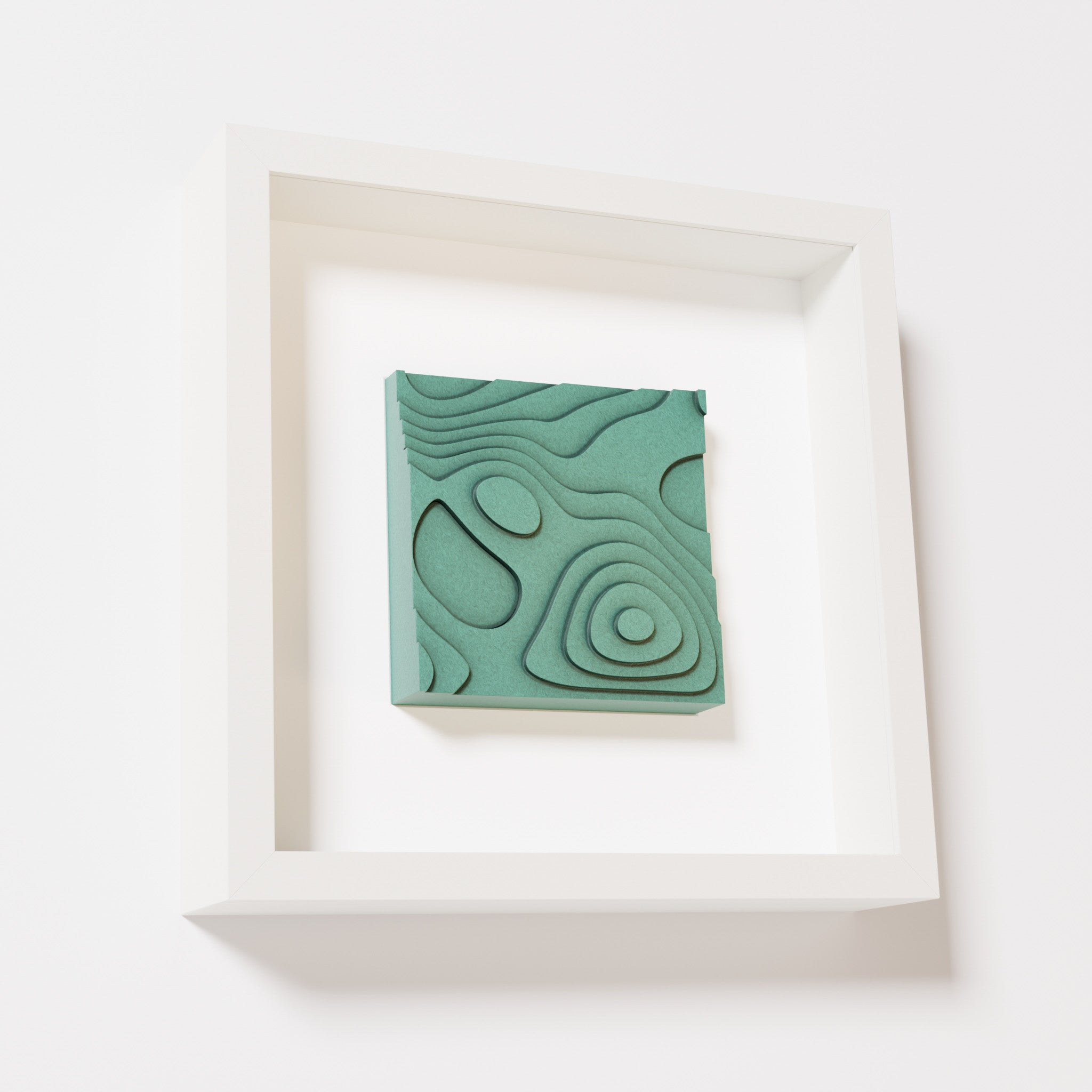 A close-up of a white shadowbox frame containing an old patina artwork depicting layered contour lines inspired by geographic elevation levels, showcasing intricate and geometric details with a textured green finish.