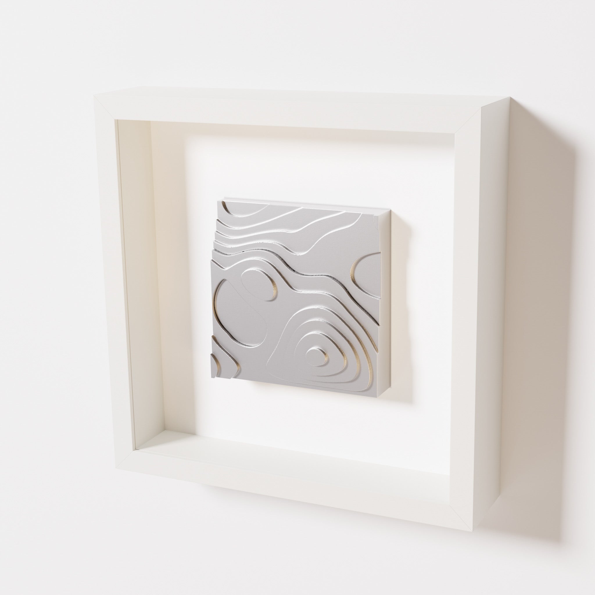 A close-up of a white shadowbox frame containing a chrome silver artwork depicting layered contour lines inspired by geographic elevation levels, showcasing reflective silver details.