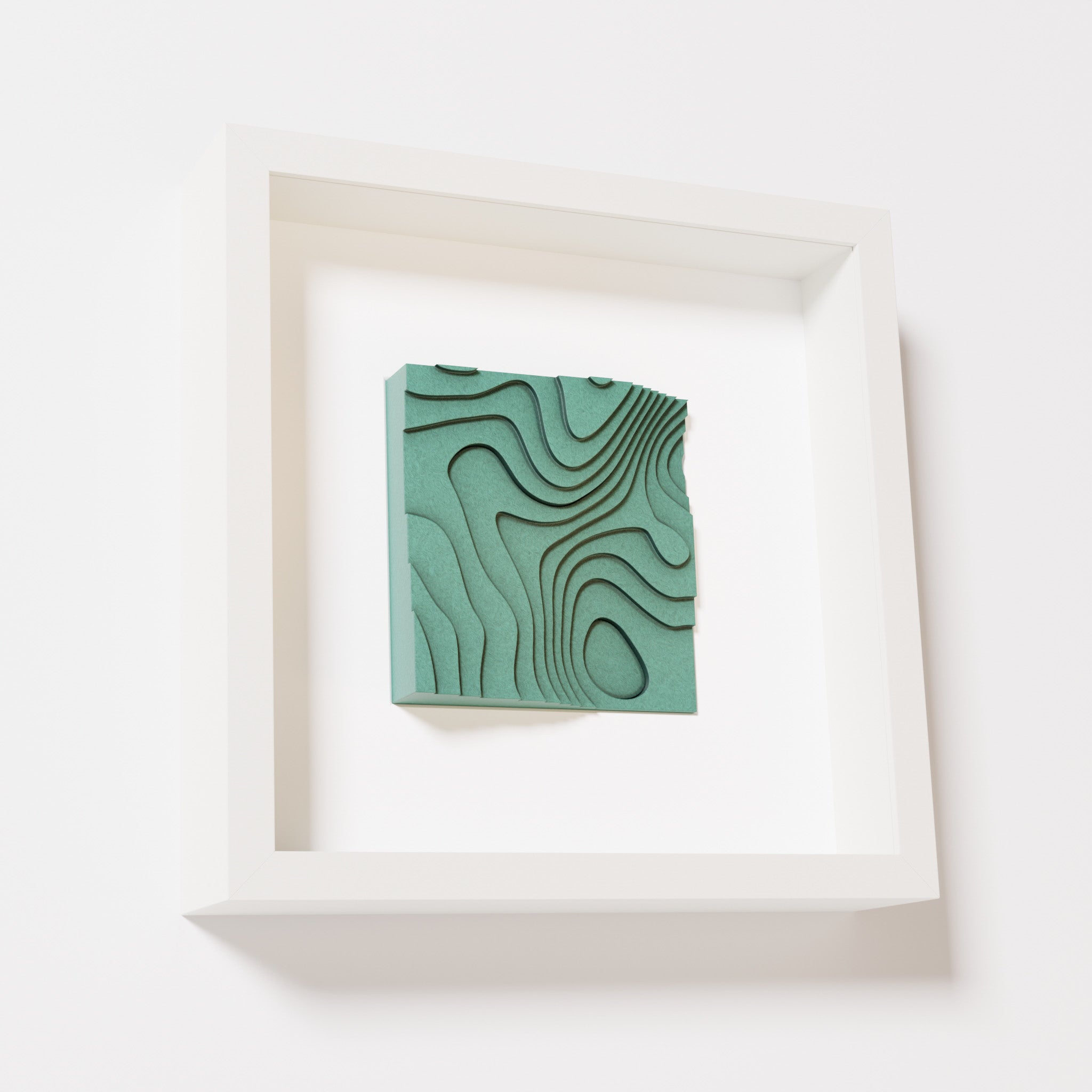 A close-up of a white shadowbox frame containing an old patina artwork depicting flowing contour lines inspired by geographic elevation levels, showcasing intricate geometric details with a textured green finish.