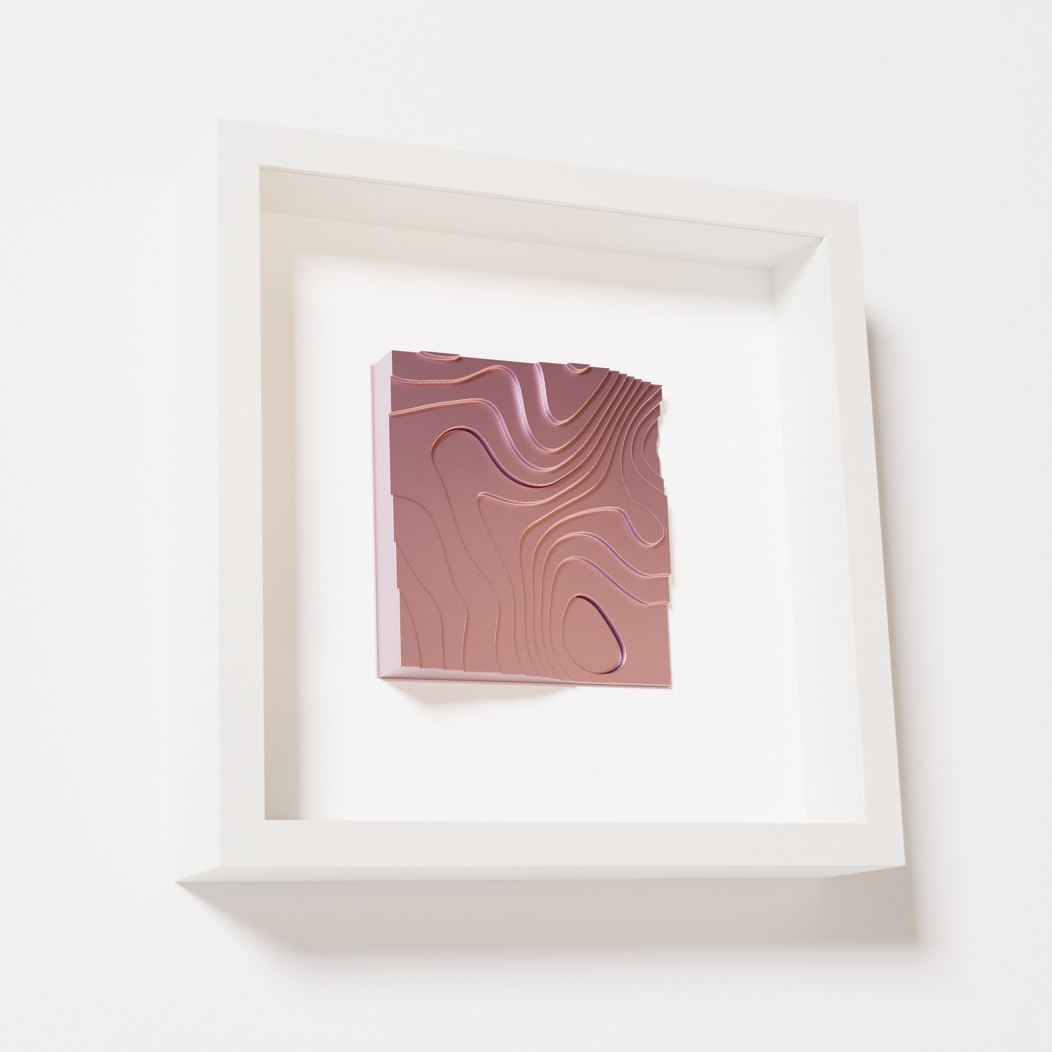 A close-up of a white shadowbox frame containing a metallic rose artwork depicting flowing contour lines inspired by geographic elevation levels, showcasing reflective pink details.