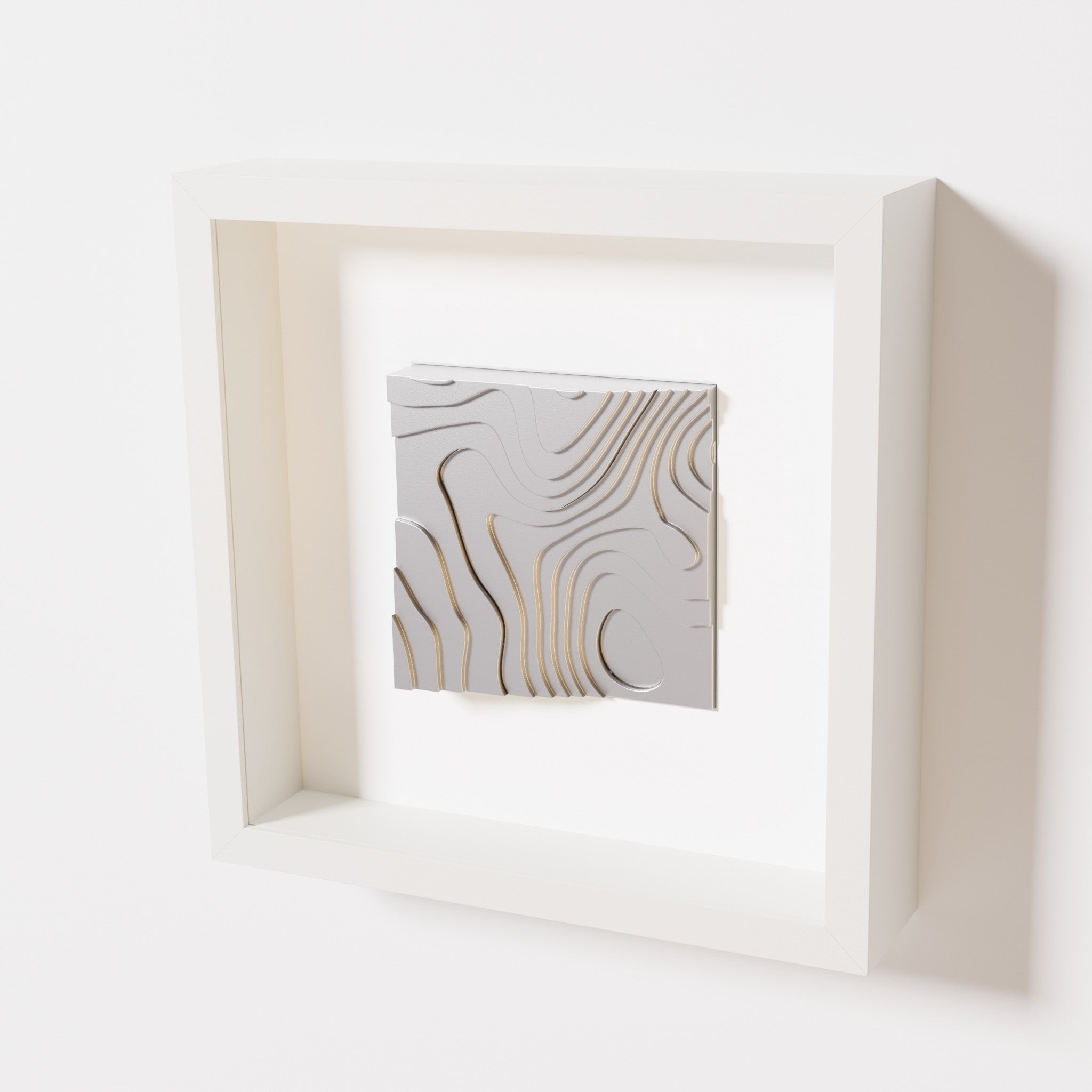 A close-up of a white shadowbox frame containing a chrome silver artwork depicting flowing contour lines inspired by geographic elevation levels, showcasing reflective silver details.