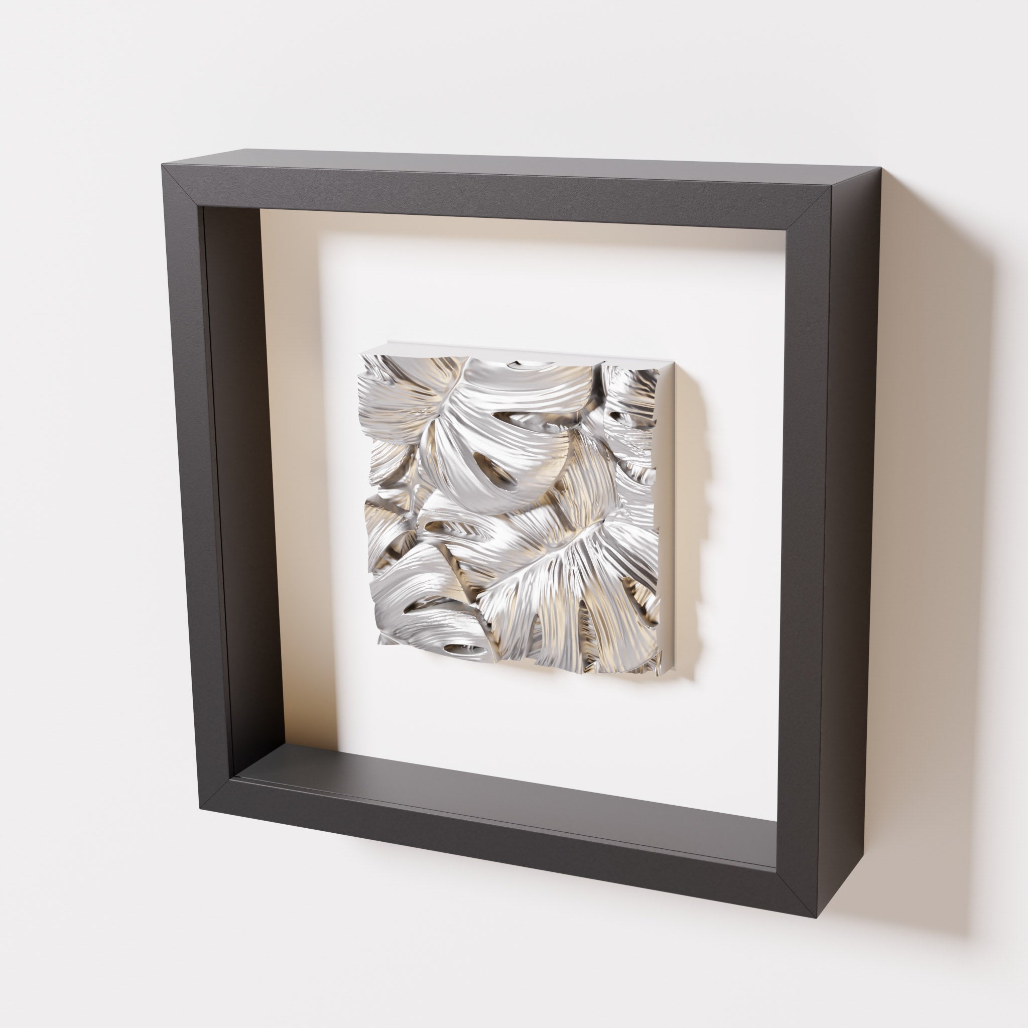 A close-up of a black shadowbox frame containing a chrome silver artwork featuring layered Monstera leaves, showcasing reflective silver highlights and intricate overlapping details.