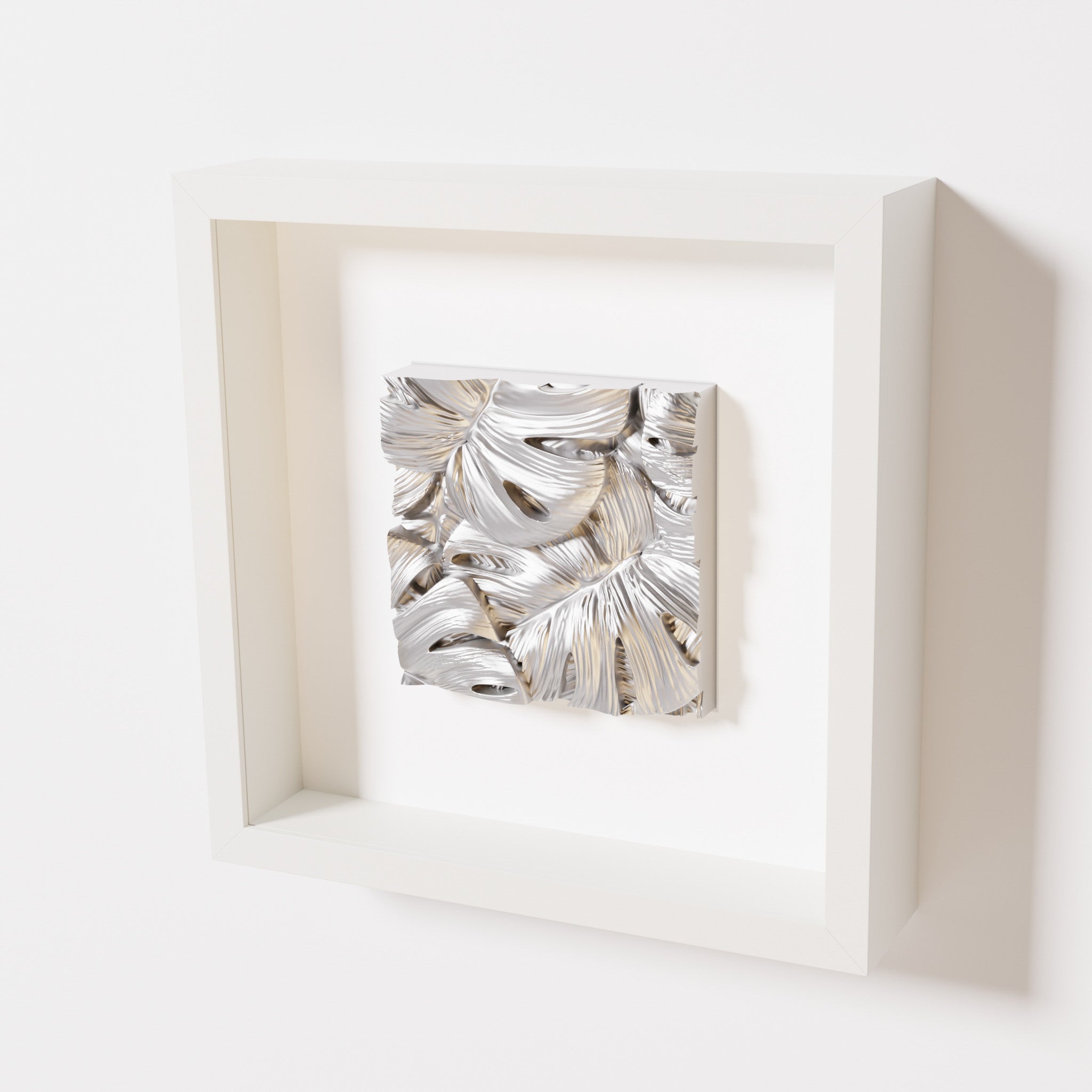 A close-up of a white shadowbox frame containing a chrome silver artwork featuring layered Monstera leaves, showcasing reflective silver highlights and intricate overlapping details.