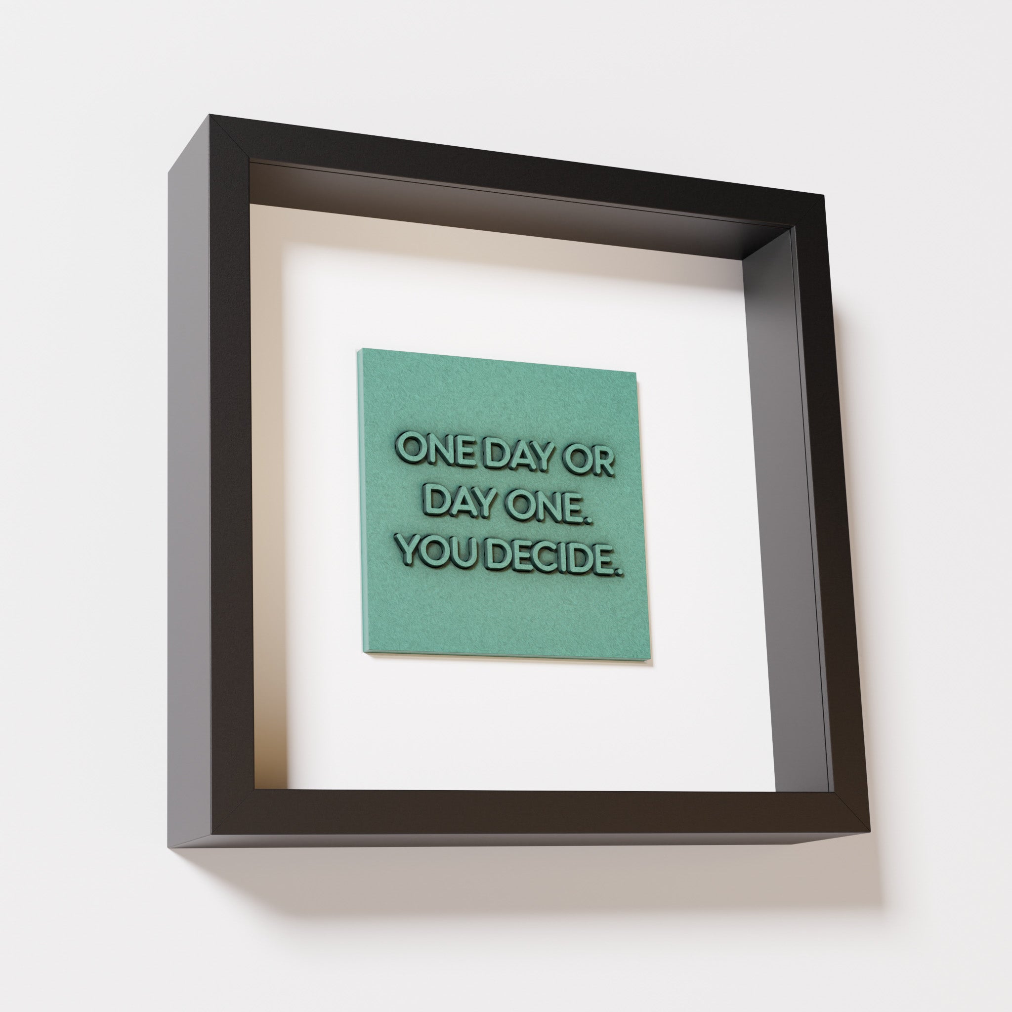 A close-up of a black shadowbox frame containing an old patina artwork featuring the phrase 'One day or day one. You decide,' with a textured teal-green finish and intricate details.