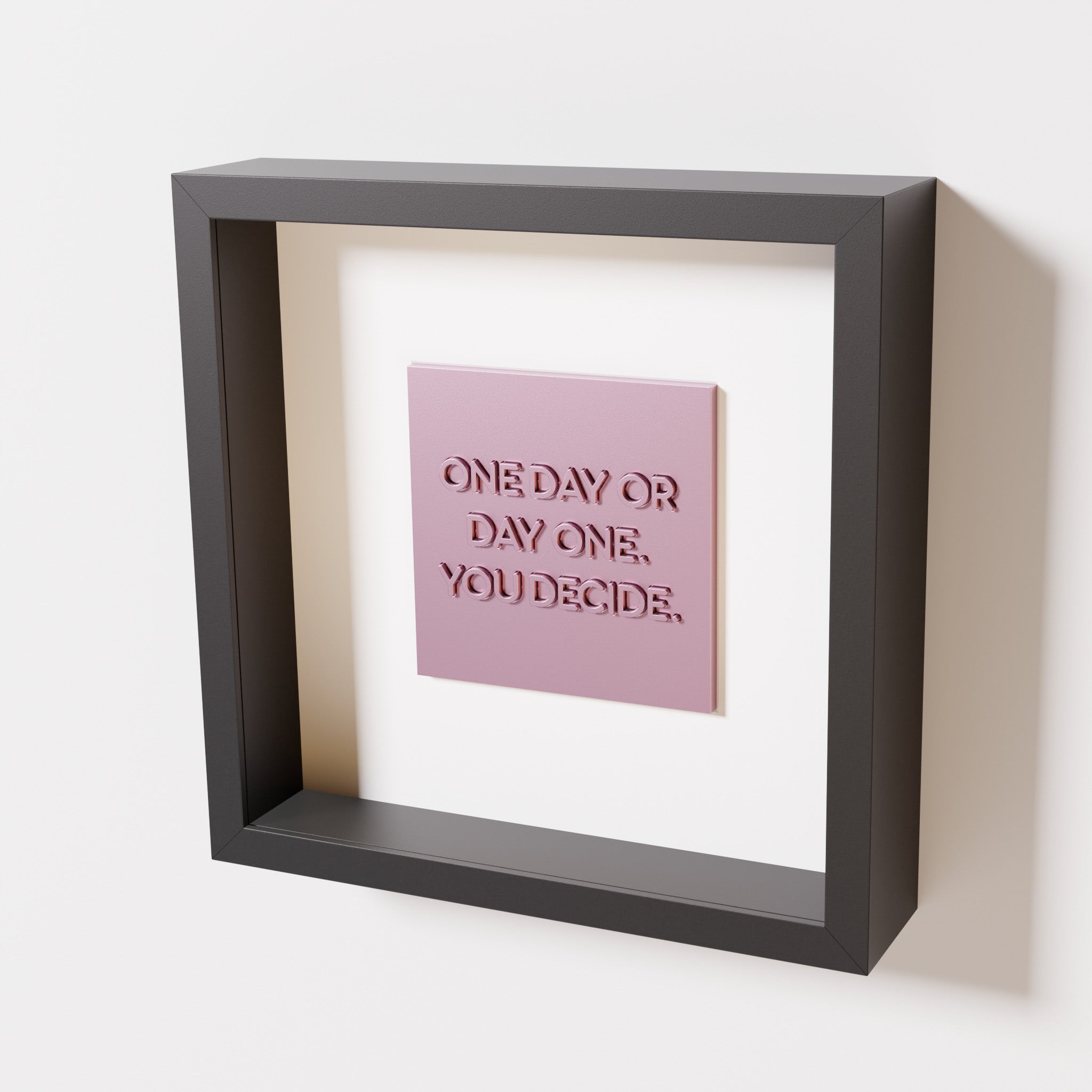 A close-up of a black shadowbox frame containing a metallic rose artwork featuring the phrase 'One day or day one. You decide,' with reflective pink highlights and embossed details.