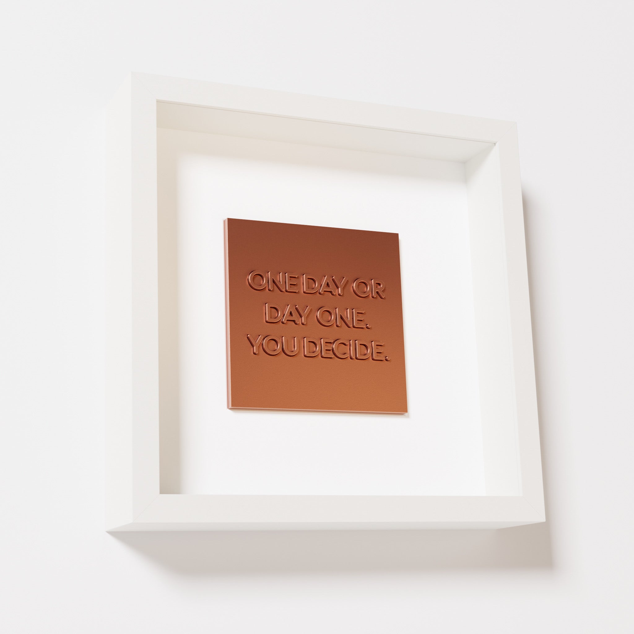 A close-up of a white shadowbox frame containing a chrome copper artwork featuring the phrase 'One day or day one. You decide,' with reflective copper highlights and embossed details.