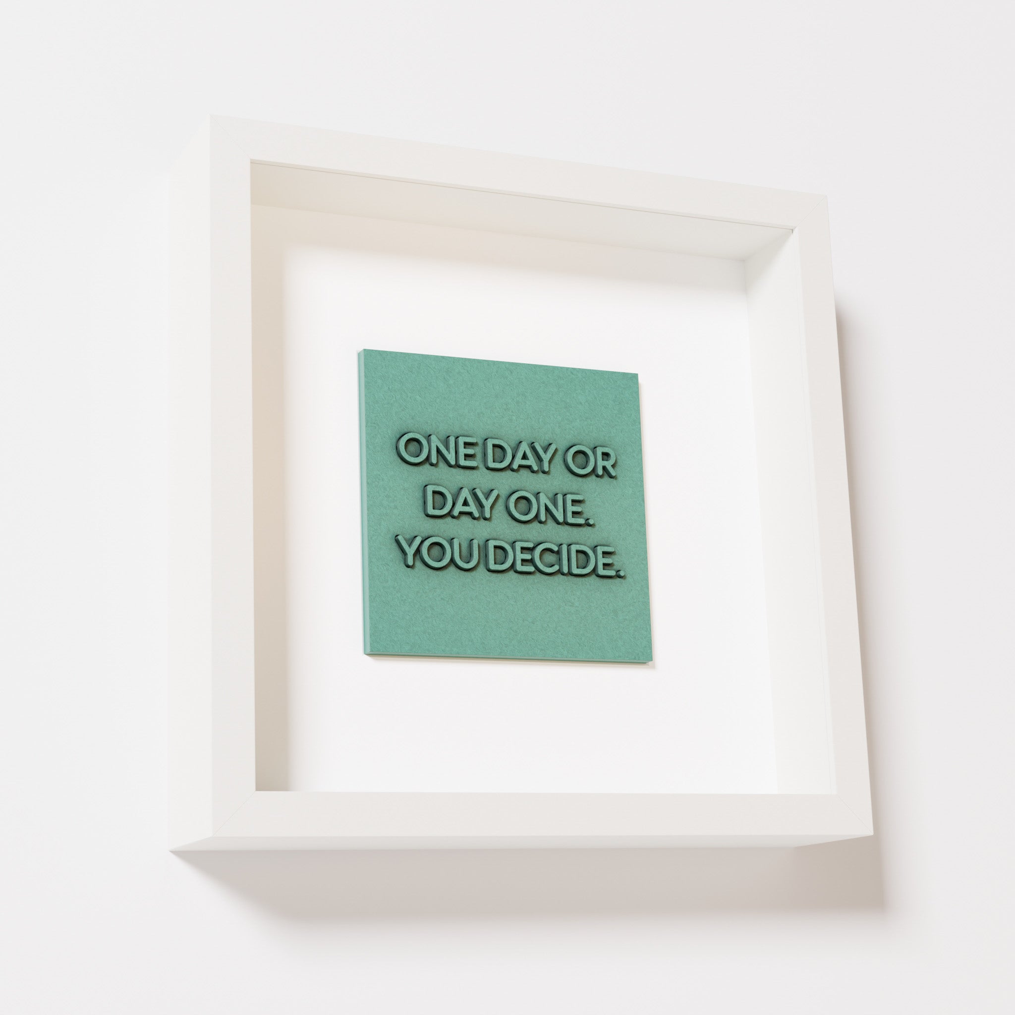 A close-up of a white shadowbox frame containing an old patina artwork featuring the phrase 'One day or day one. You decide,' with a textured teal-green finish and intricate details.